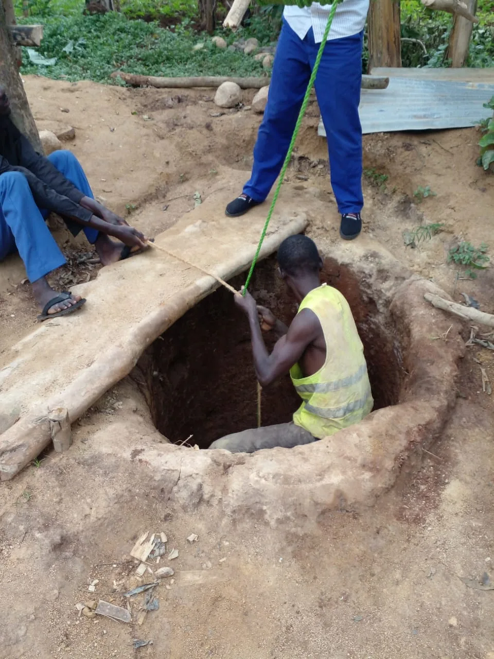 Adults using a rope while a worker stands inside a hand-dug water source.